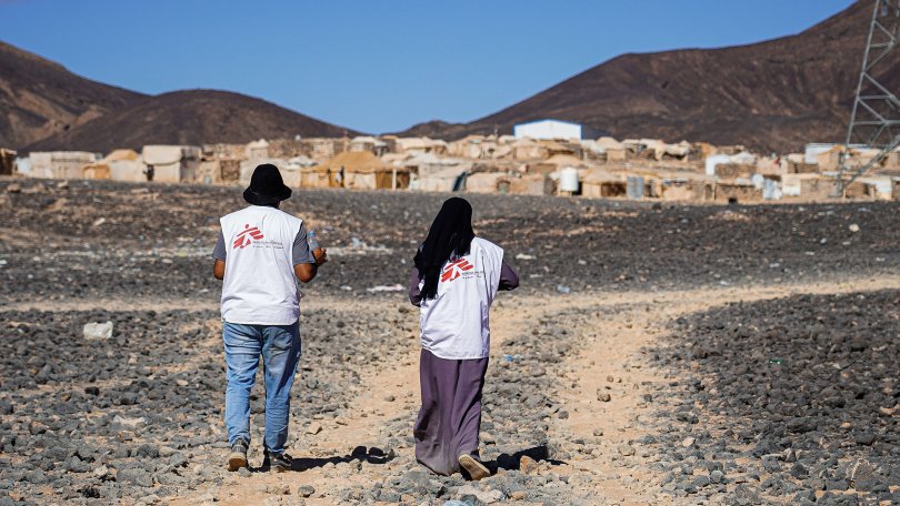 Two people walking down a dirt road with mountains in the background.
