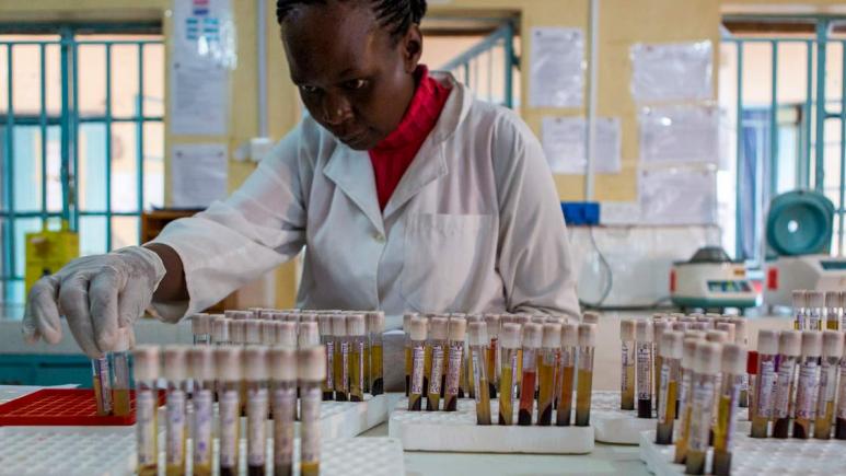 A woman is working in a lab, carefully handling numerous test tubes at the epicentre.